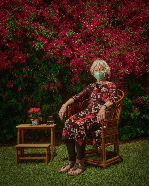 Elderly woman wearing a face mask sits in a wicker chair in a garden, with a small table and flowers nearby, and lush bougainvillea in the background.